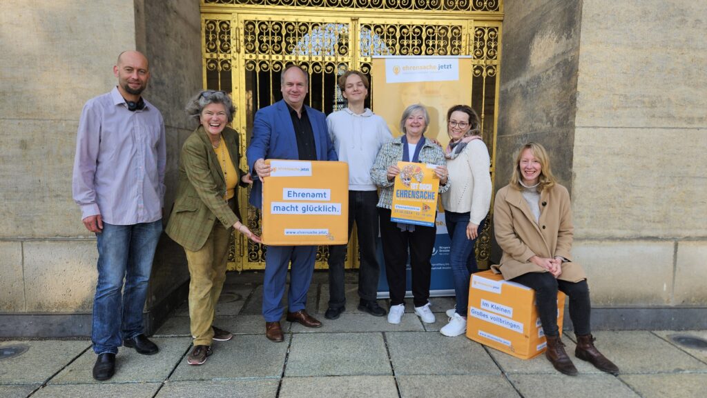 Menschen stehen vor der Goldenen Pforte des Dresdner Rathauses. Sie halten ein Schild: Ehrenamt macht glücklich.