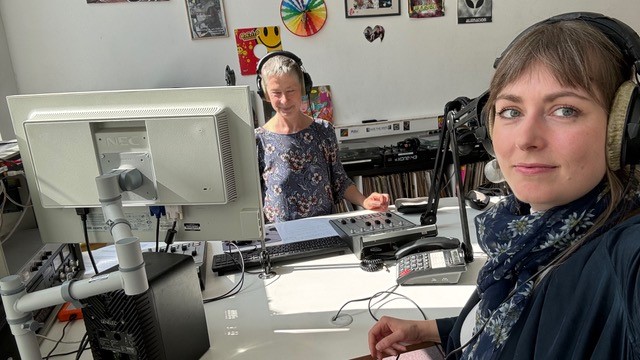 Im Vordergrund eine Frau mit Kopfhörern. Zu sehen ist ein Radiostudio. Am Tisch steht ihr eine zweite Moderatorin gegenüber.