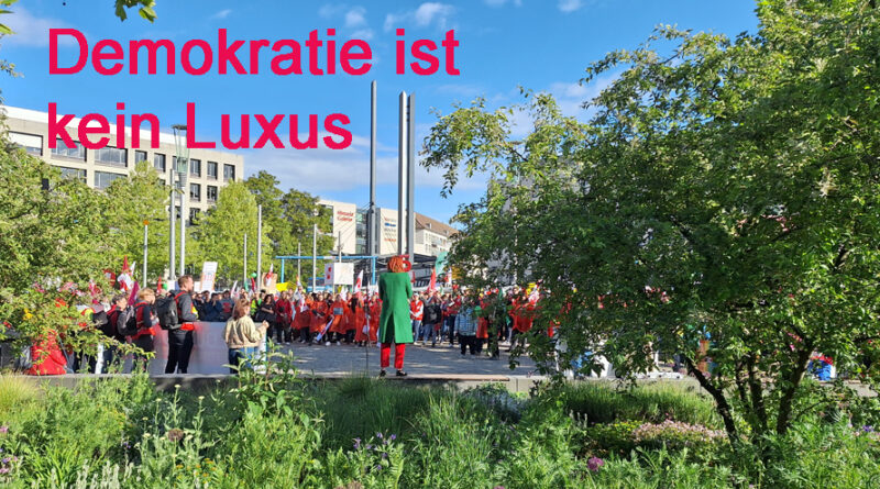 Menschen auf dem Postplatz. Eine Rednerin spricht. Darüber steht: Demokratie ist kein Luxus.