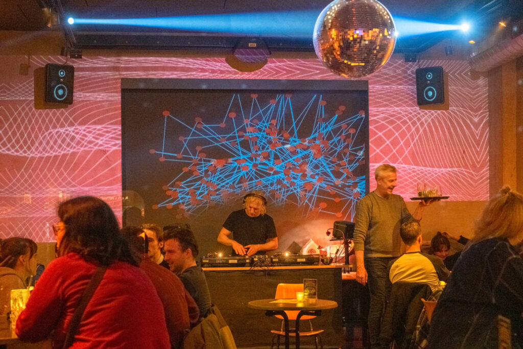 Das Bild zeigt einen Raum in dem links und rechts Menschen an einem Tisch sitzen. Ein Kellner bringt etwas zu trinken. In der Mitte an der Hinterwand ist ein DJ der auflegt.