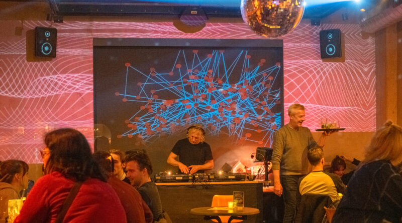 Das Bild zeigt einen Raum in dem links und rechts Menschen an einem Tisch sitzen. Ein Kellner bringt etwas zu trinken. In der Mitte an der Hinterwand ist ein DJ der auflegt.