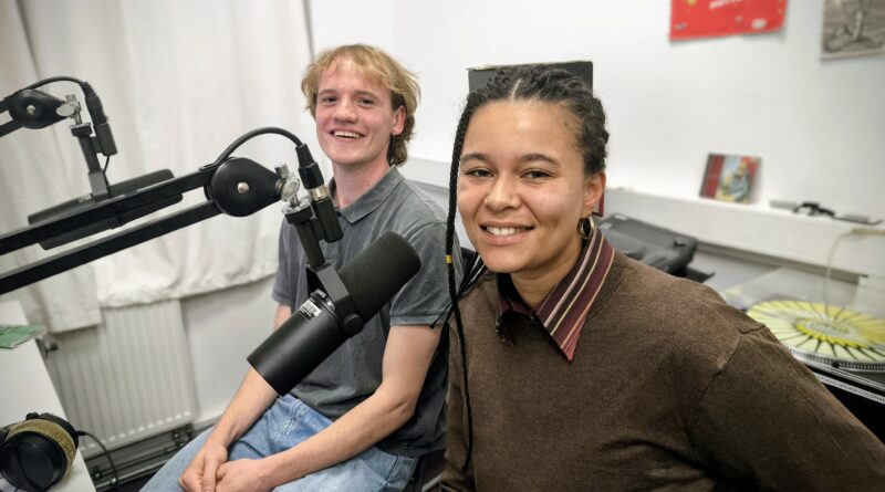 Das Bild zeigt 2 Menschen die im Aufnahmestudio sitzen und Mikrophone vor ihrem Gesicht haben.