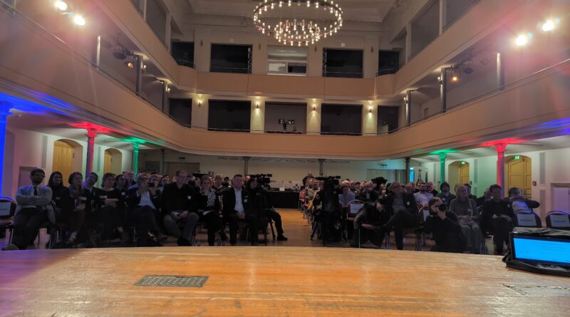 Das Bild zeigt einen großen Saal von der Bühne aus. Viele Menschen sitzen auf Stühlen und sind schick angezogen.