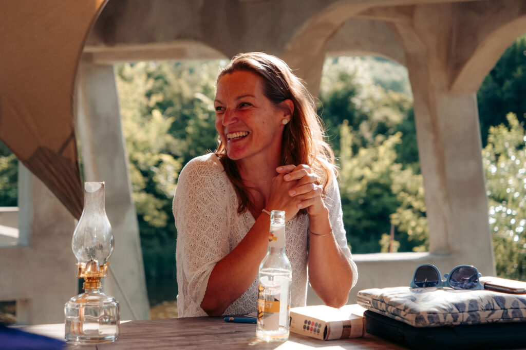 Das Bild zeigt eine lachende Frau die an einem Tisch sitzt. Auf dem Tisch vor ihr steht eine leere Limoflasche.