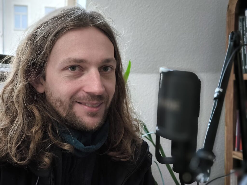 Das Bild zeigt ein Mann mit langem Haar, der vor Rauhfasertapete sitzt.