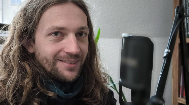 Das Bild zeigt ein Mann mit langem Haar, der vor Rauhfasertapete sitzt.