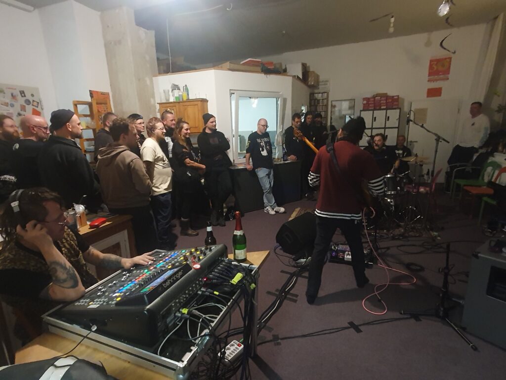 Das Bild zeigt einen kleinen Raum mit ca 20 Menschen. Diese stehen um eine Band herum die spielt. Links im Bild ist ein Tontechniker vor einem Mischpult.