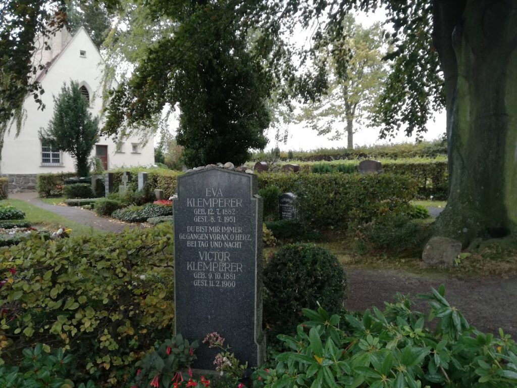 Das Bild zeigt einen Grabstein auf einem Friedhof. Um den Grabstein stehen viele Pflanzen.