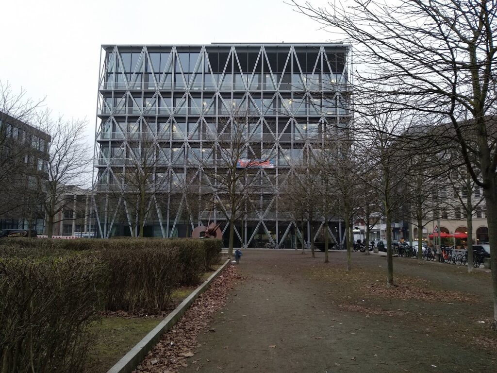 Das Bild zeigt ein Haus in Berlin. Es ist kein Häuserblock sonder steht gesondert neben einem kleinen Park mit wenig Pflanzen.