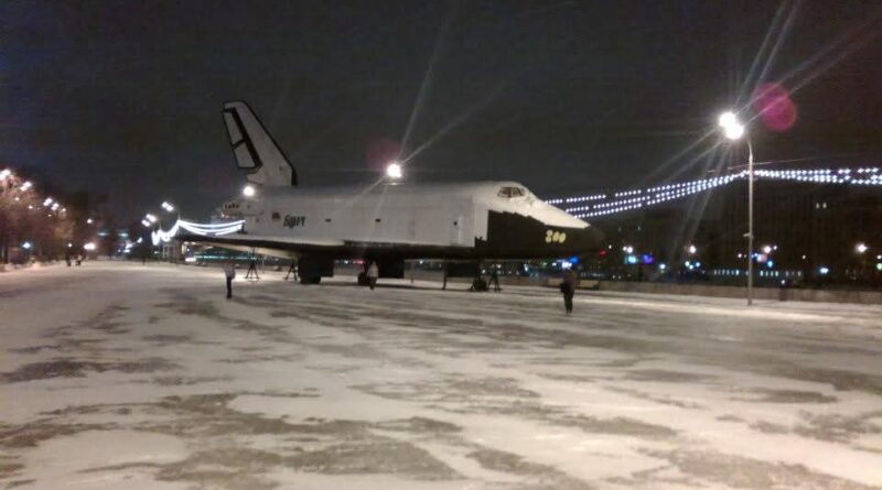 Das Bild zeigt das Spaceshuttle Buran im Winter bei Nacht in Moskau. Das Spaceshuttle steht auf einer breiten leeren Straße in der Stadt. Im Hintergrund sind die Lichter der Häuser zu sehen.