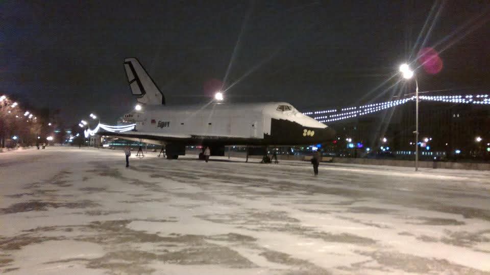 Das Bild zeigt das Spaceshuttle Buran im Winter bei Nacht in Moskau. Das Spaceshuttle steht auf einer breiten leeren Straße in der Stadt. Im Hintergrund sind die Lichter der Häuser zu sehen.
