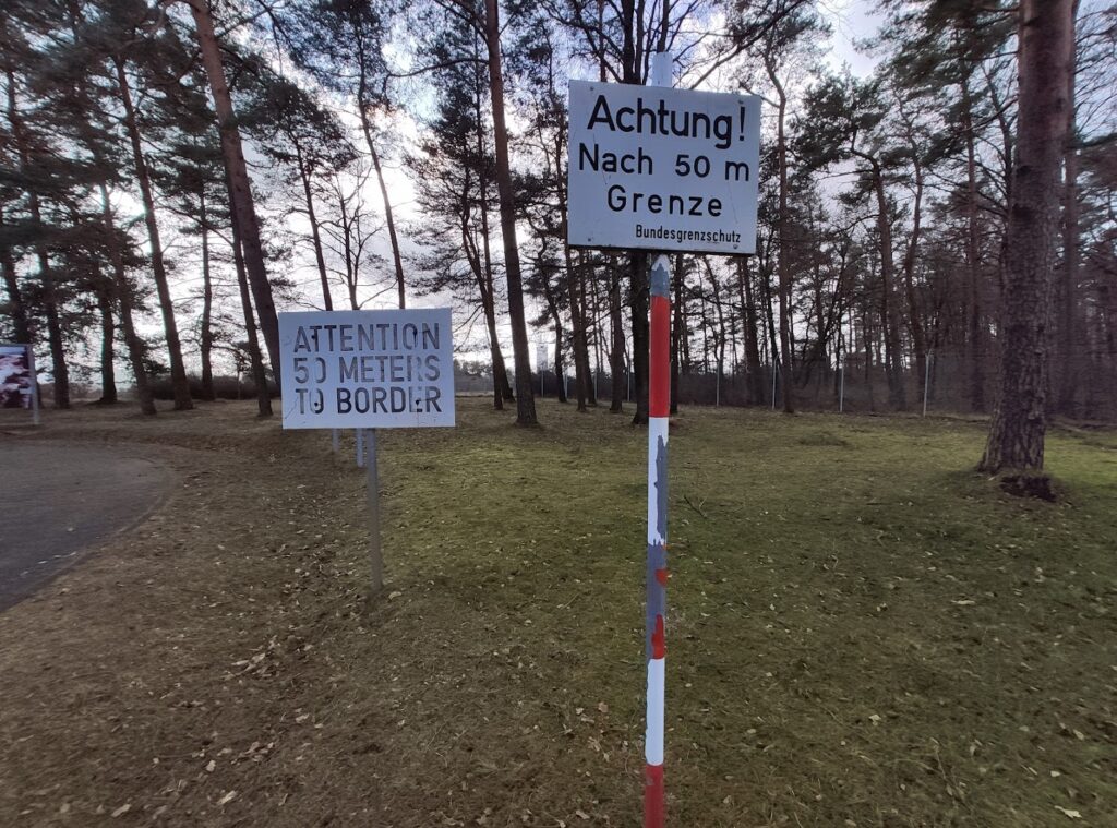 Das Bild zeigt ein Waldstück bei Tag. Zwei Schilder stehen im Vordergrund. Auf den Schildern steht: Achtung! Nach 50 m Grenze. Und auf dem anderen dasselbe in Englisch.