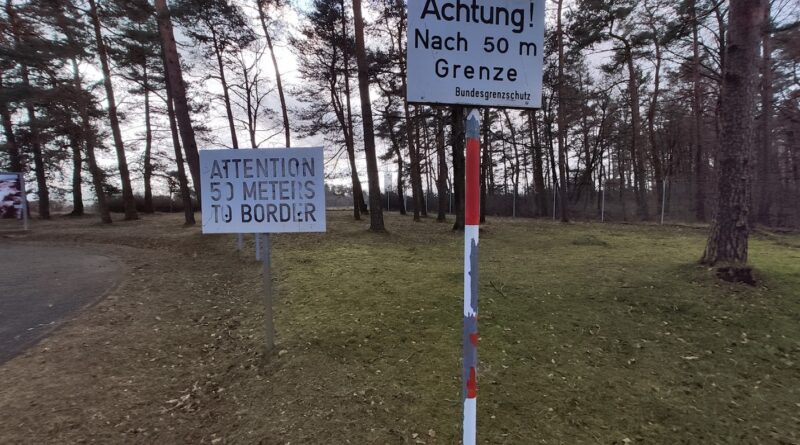 Das Bild zeigt ein Waldstück bei Tag. Zwei Schilder stehen im Vordergrund. Auf den Schildern steht: Achtung! Nach 50 m Grenze. Und auf dem anderen dasselbe in Englisch.