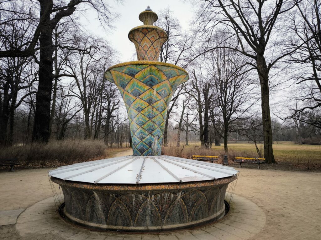 Das Bild zeigt den Mosaikbrunnen im Großen Garten im sehr frühen Frühling. Der Brunnen ist ca 3 m hoch und hat mandalaartige Muster die seine Brunnensäule verzieren. Neben dem Brunnen stehen Bäume die keine Blätter tragen.