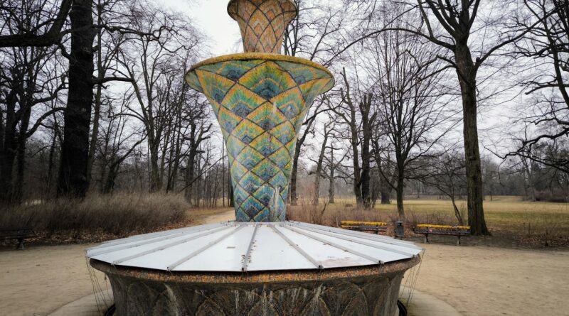 Das Bild zeigt den Mosaikbrunnen im Großen Garten im sehr frühen Frühling. Der Brunnen ist ca 3 m hoch und hat mandalaartige Muster die seine Brunnensäule verzieren. Neben dem Brunnen stehen Bäume die keine Blätter tragen.