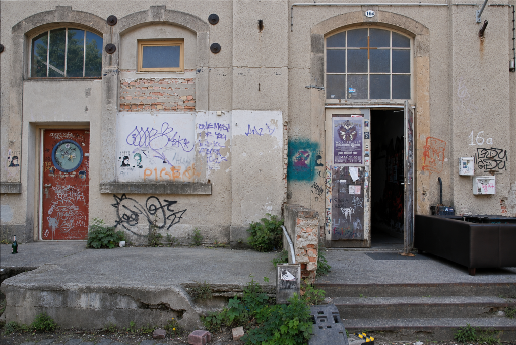 Das Bild zeigt eine verfallene Hauswand mit Grafittis drauf und eine offene Metalltür.