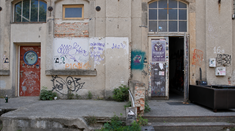 Das Bild zeigt eine verfallene Hauswand mit Grafittis drauf und eine offene Metalltür.