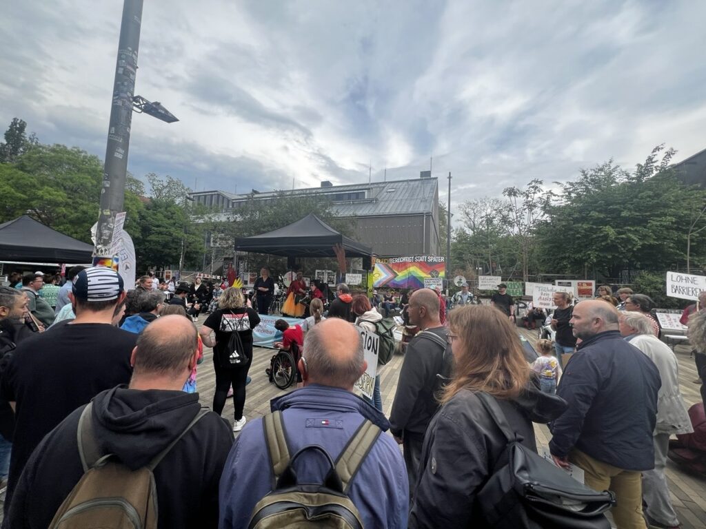Viele Menschen stehen auf einem Platz. Einige von ihnen halten Banner hoch. Auf den steht: Gleichberechtigung für alle und Menschenrechte sind nicht verhandelbar. In der Mitte steht ein Pavillon, dort spricht Manuel Schramm.