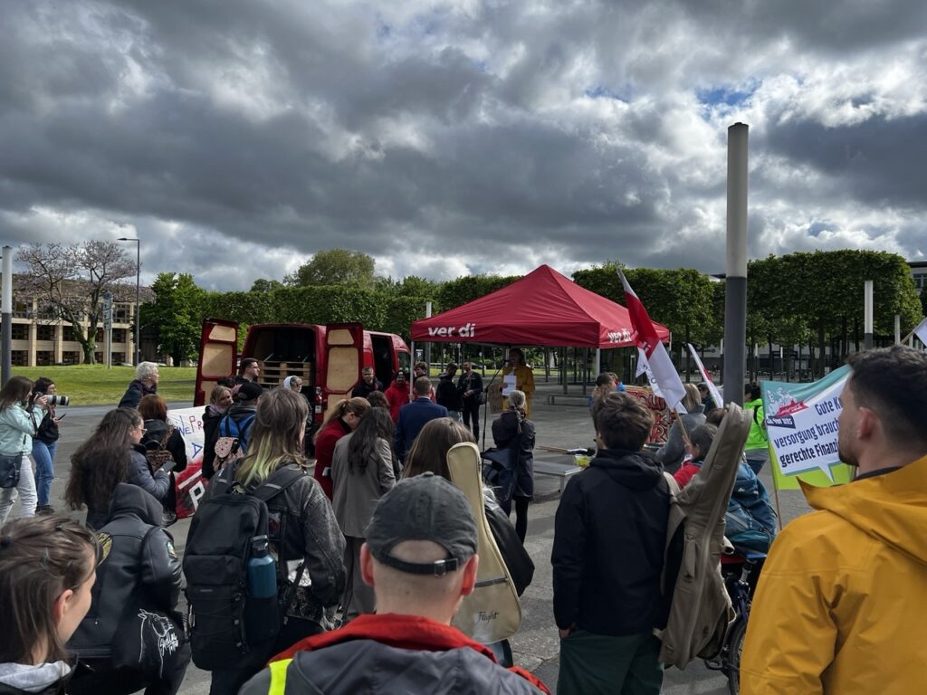 Auf dem Bild sind mehrere Menschen die vor einem roten Pavillon stehen. Auf dem Pavillon steht ver.di. Sie halten Banner hoch, auf einem steht: Versorgung braucht gerechte Finanzierung.