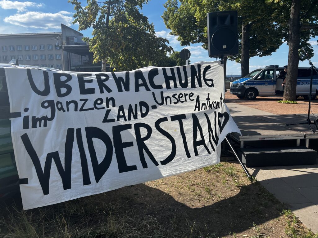 Im Vordergrun dist ein Banner it der Aufschrift: Überwachung im ganzen Land unsere Antwort Widerstand. Im Hintergrund ist ein Polizeiauto zu sehen.