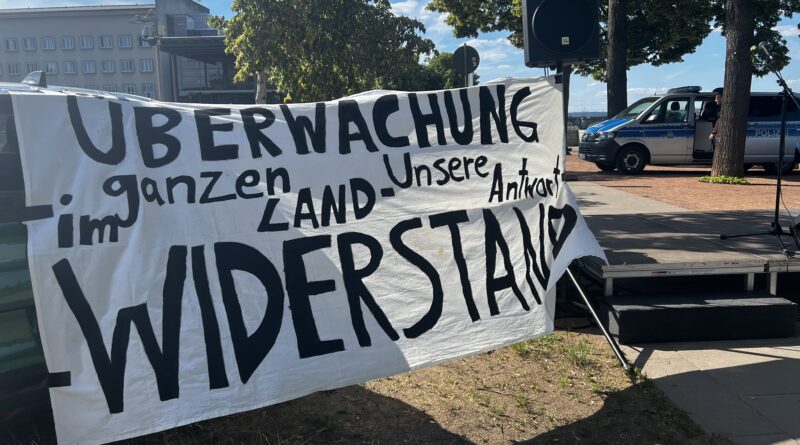 Im Vordergrun dist ein Banner it der Aufschrift: Überwachung im ganzen Land unsere Antwort Widerstand. Im Hintergrund ist ein Polizeiauto zu sehen.